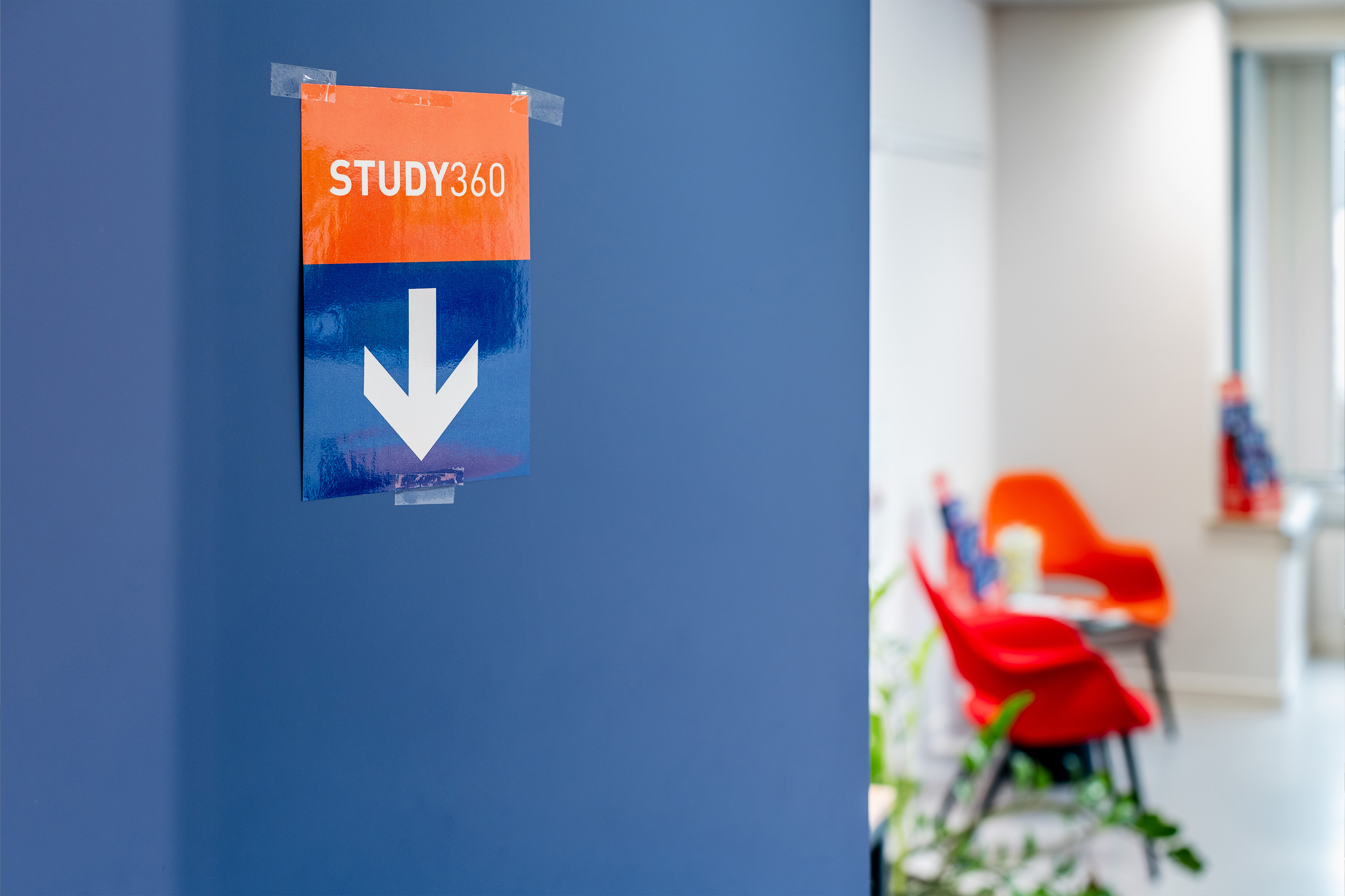 Zicht in een gang met in de verte 2 oranje stoelen. Links op de voorgrond een deel van een muur met signalisatie van STUDY360 op