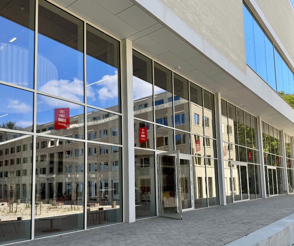 Buitenaanzicht van de voorzijde van Bavet met de hoge glasramen en weerspiegeling van lucht en gebouwen aan de overzijde in.