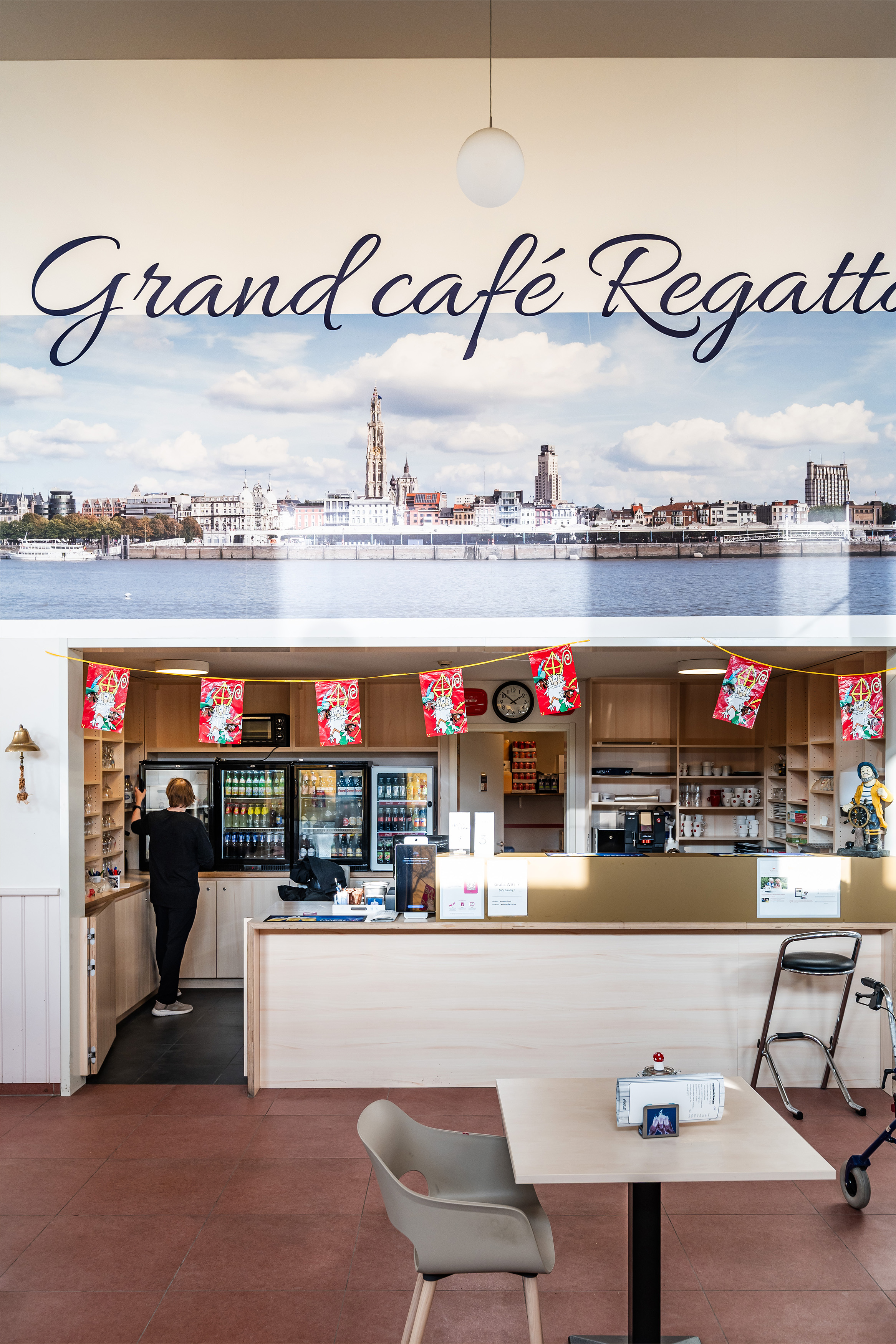 frontaal zicht op de koffiebar met de toog, gevulde ijskasten en gezellige aankleding in de vorm van vlaggetjes en een grote foto van het uitzicht op de stad Antwerpen vanaf Linkeroever