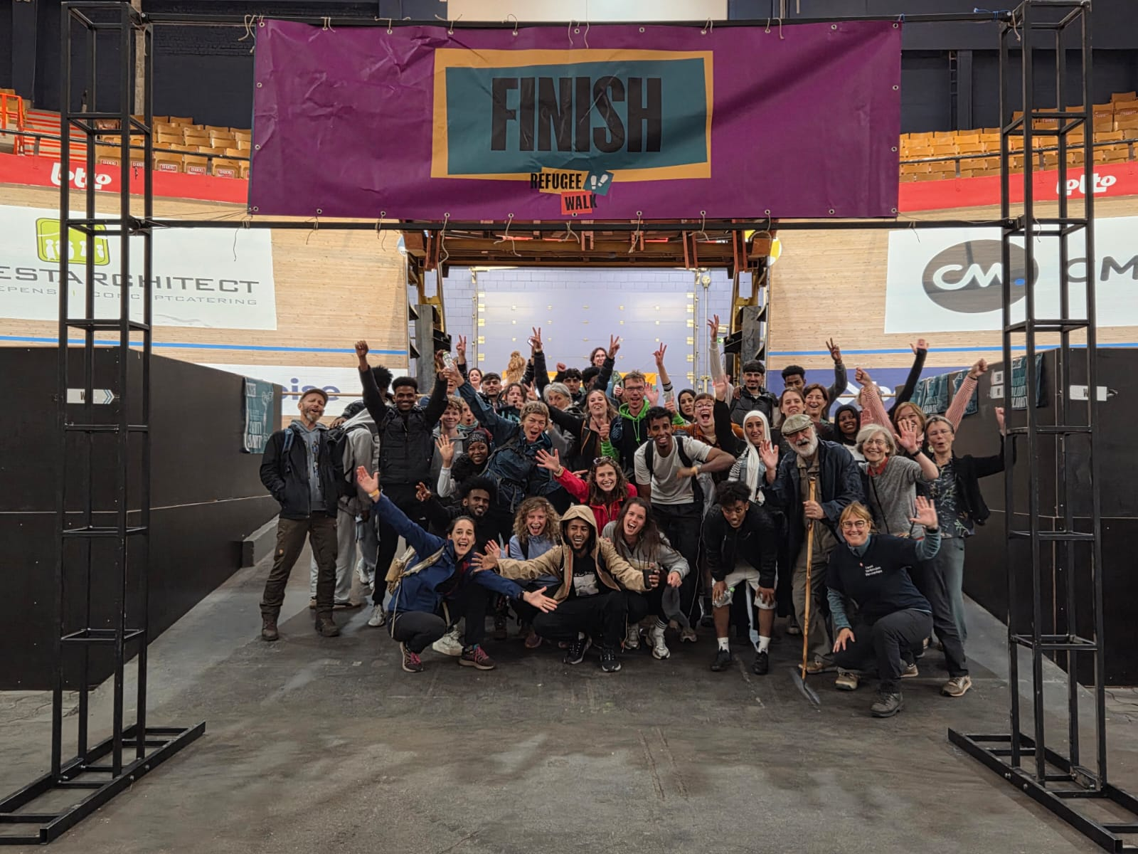 groep enthousiaste mensen onder een paarse finishboog met de tekst 'finish' na de Refugee Walk, ze juichen en poseren