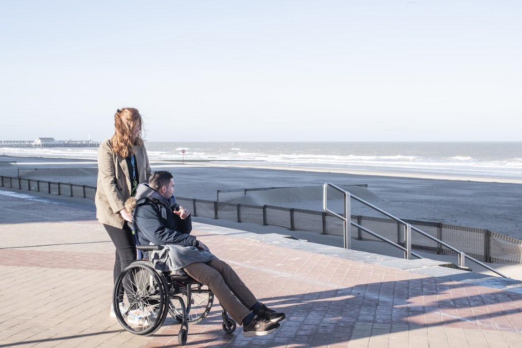 Medewerkster met jonge man in rolstoel kijken naar de zee vanop de dijk