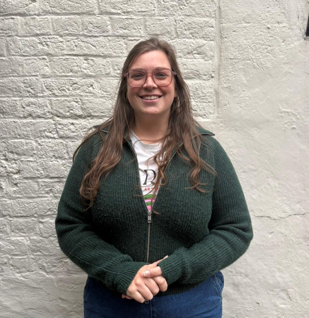 Portretfoto van Maxine Broeckaert. Een lachende, jonge vrouw met lang bruin haar en een bril.