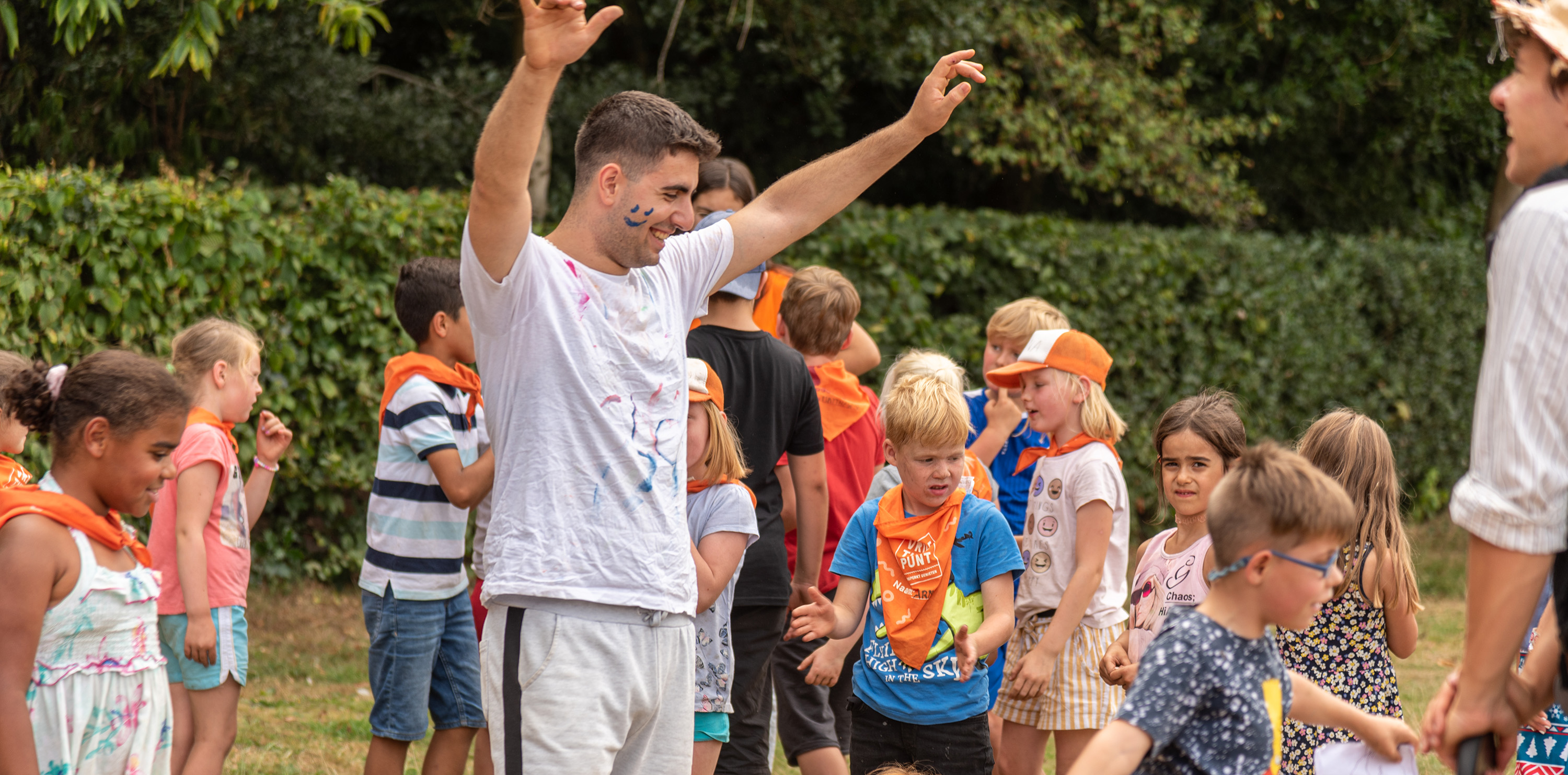 Een groep kinderen met oranje sjaaltjes speelt buiten op de speelpleinwerking naast een jonge mannelijke animator.