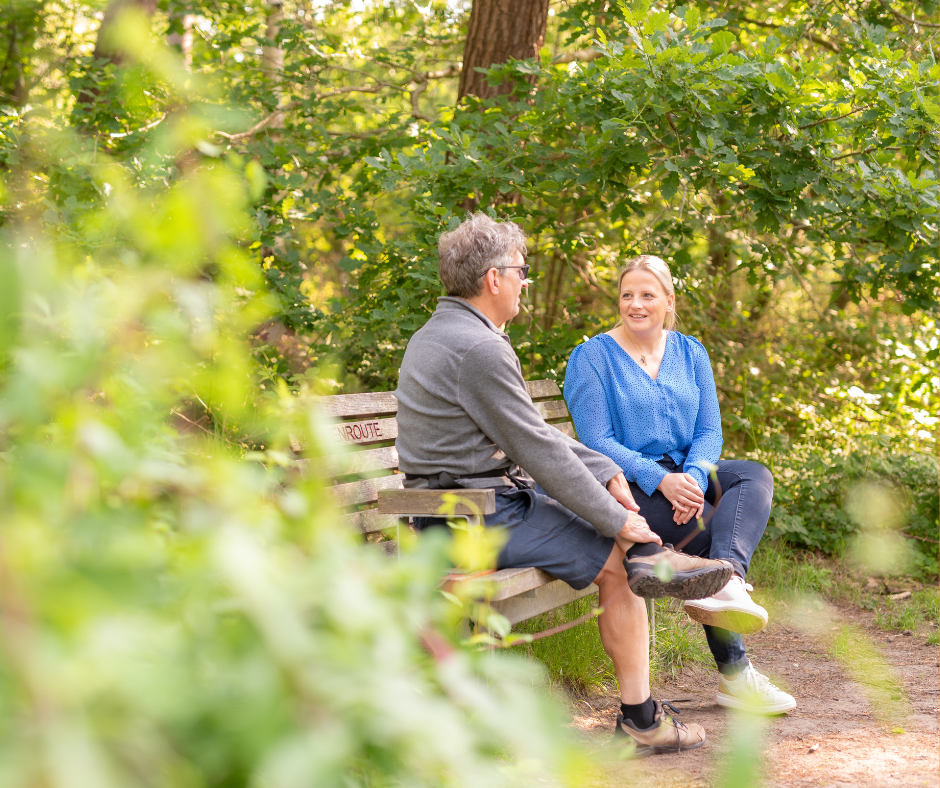 Vrouwelijke begeleidster en mannelijke cliënten zitten pratend op een bankje in de zomerse natuur.