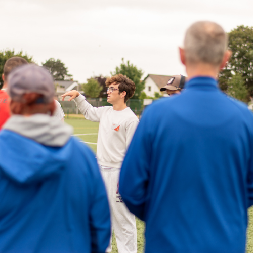 Jonge mannelijke jobstudent wijst een richting aan op een voetbalplein terwijl omstaanders kijken.