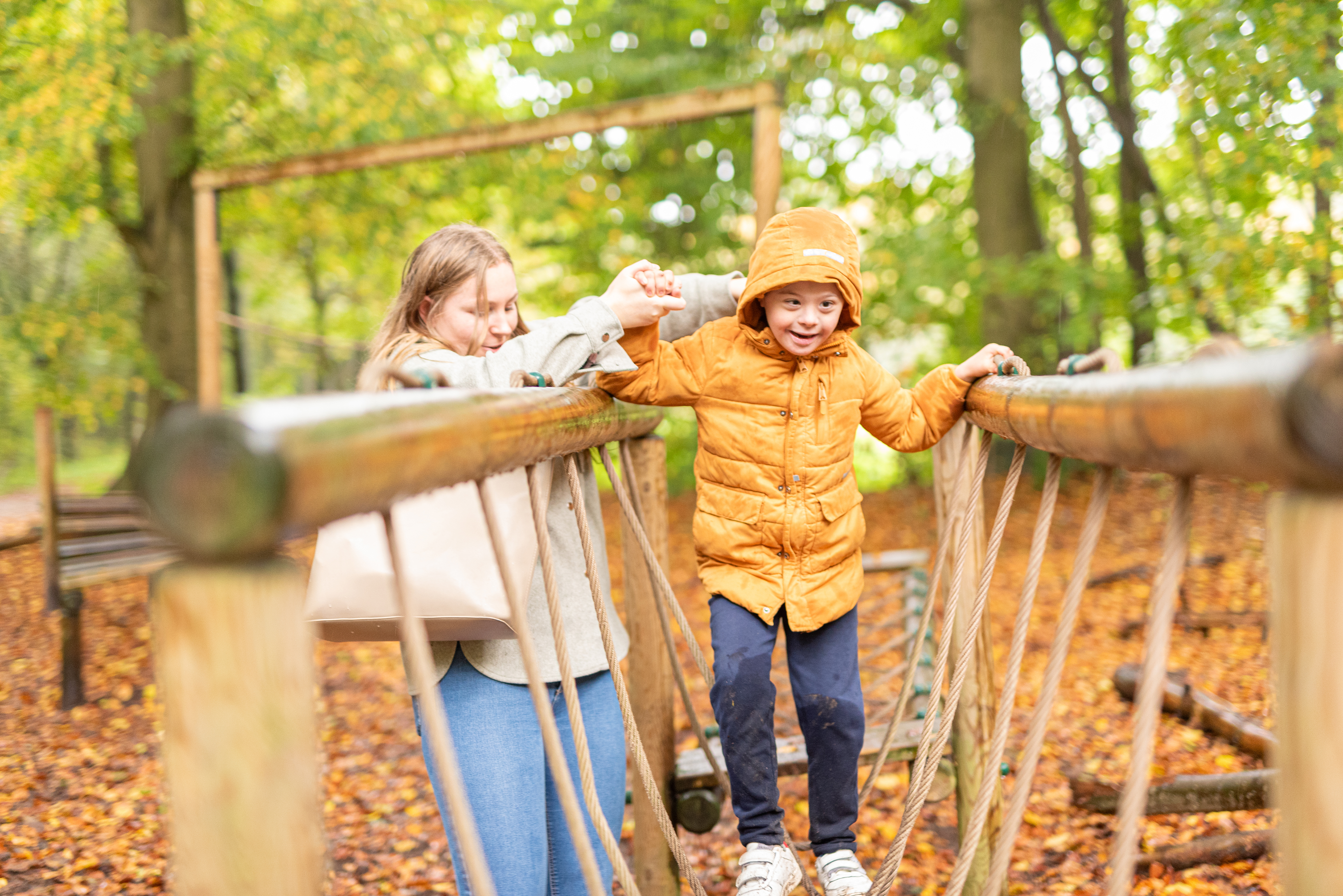 Een kind in een oranje jas loopt over een houten touwbrug in een bos. Een volwassene naast hem houdt zijn hand vast ter ondersteuning.