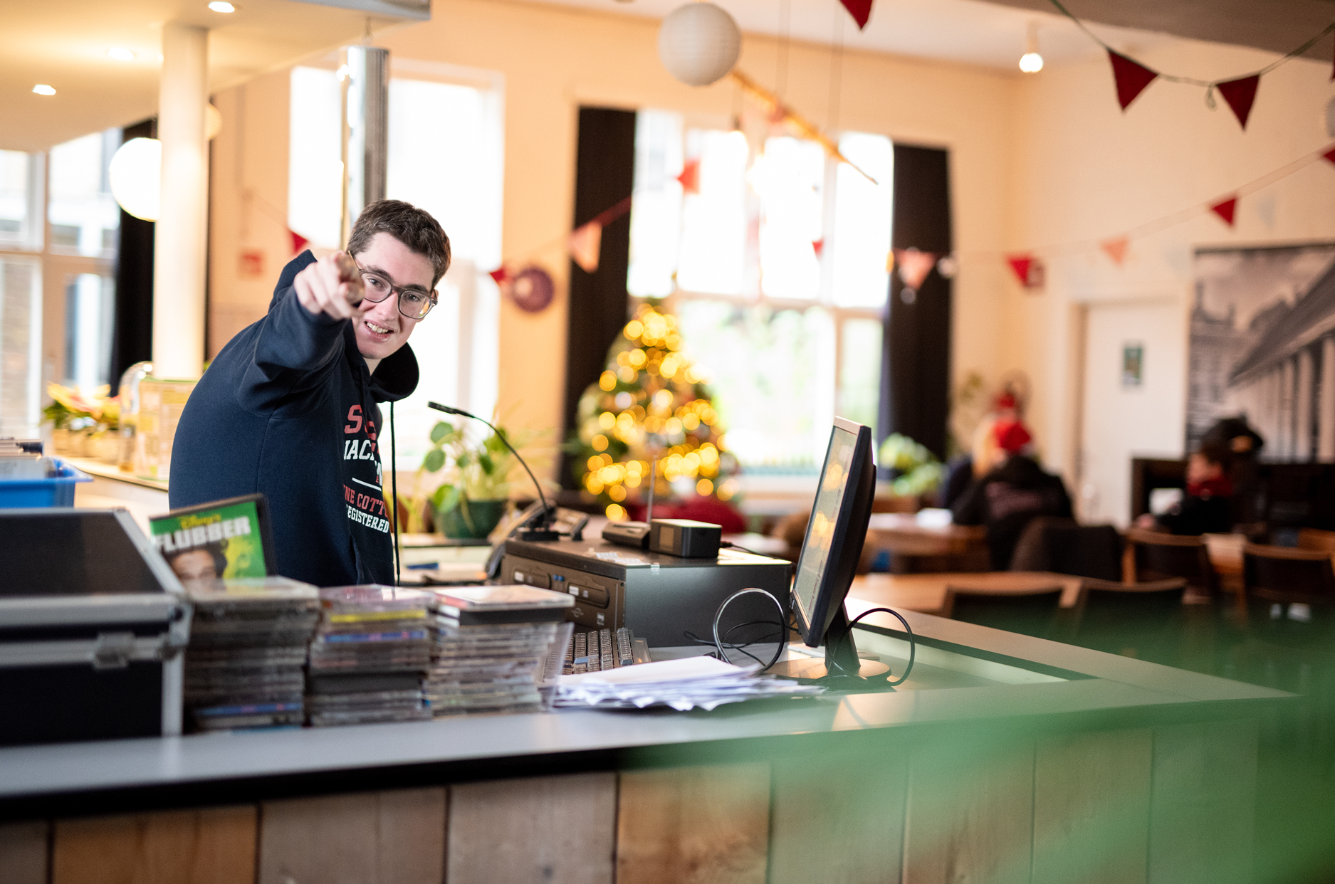Deelnemer met bril achter een balie wijst en lacht naar de camera in Café Alloo Alloo; kerstversiering op de achtergrond.