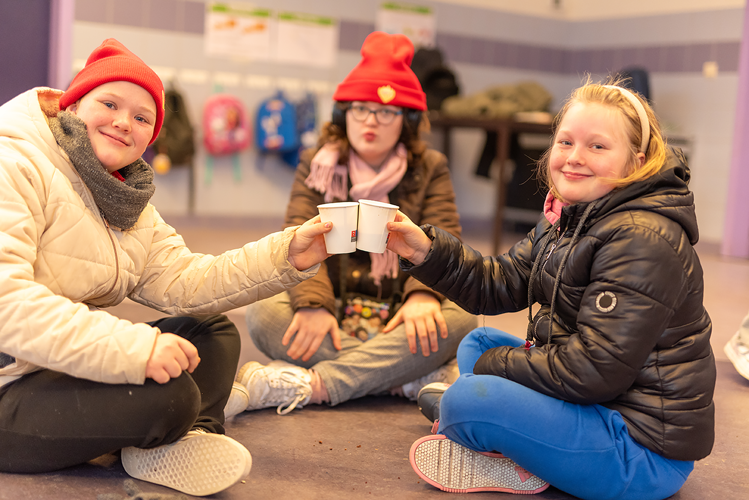 Drie kinderen zitten binnen op de grond in winterkleding en klinken vrolijk met bekertjes warme drank