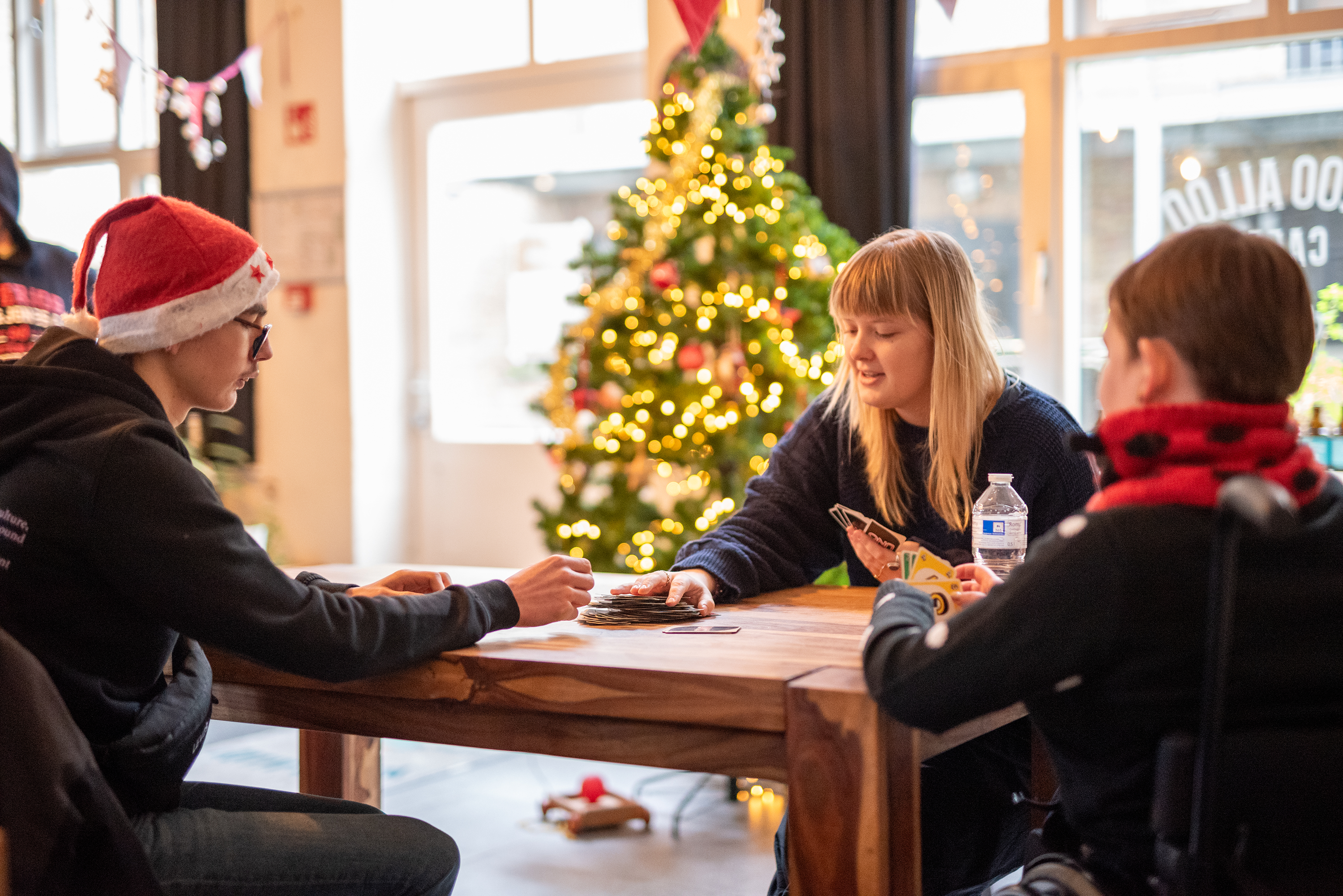 Jongvolwassenen zitten samen rond een houten tafel en spelen een kaartspel, met een verlichte kerstboom op de achtergrond.
