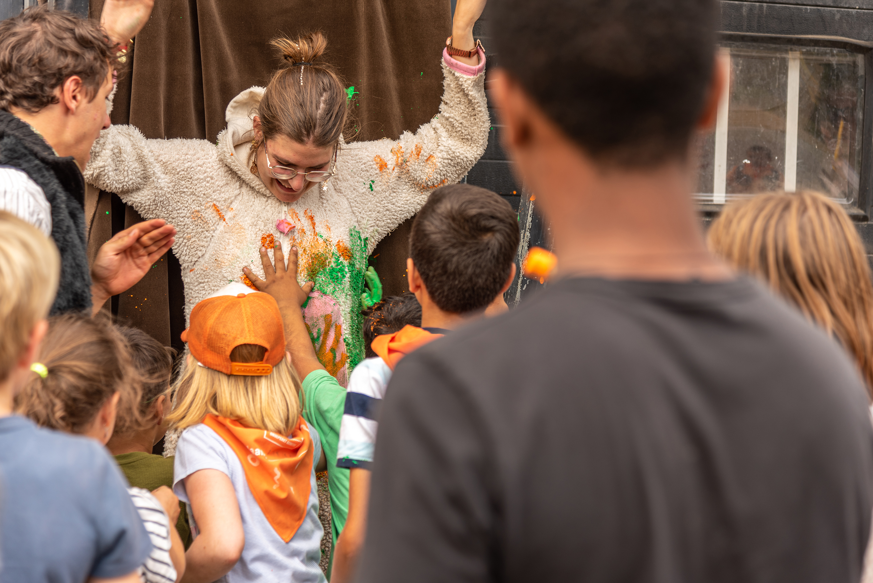 Een groep kinderen gooit kleurrijke verf op een lachende vrijwilligster met opgestoken armen tijdens een speelse activiteit.