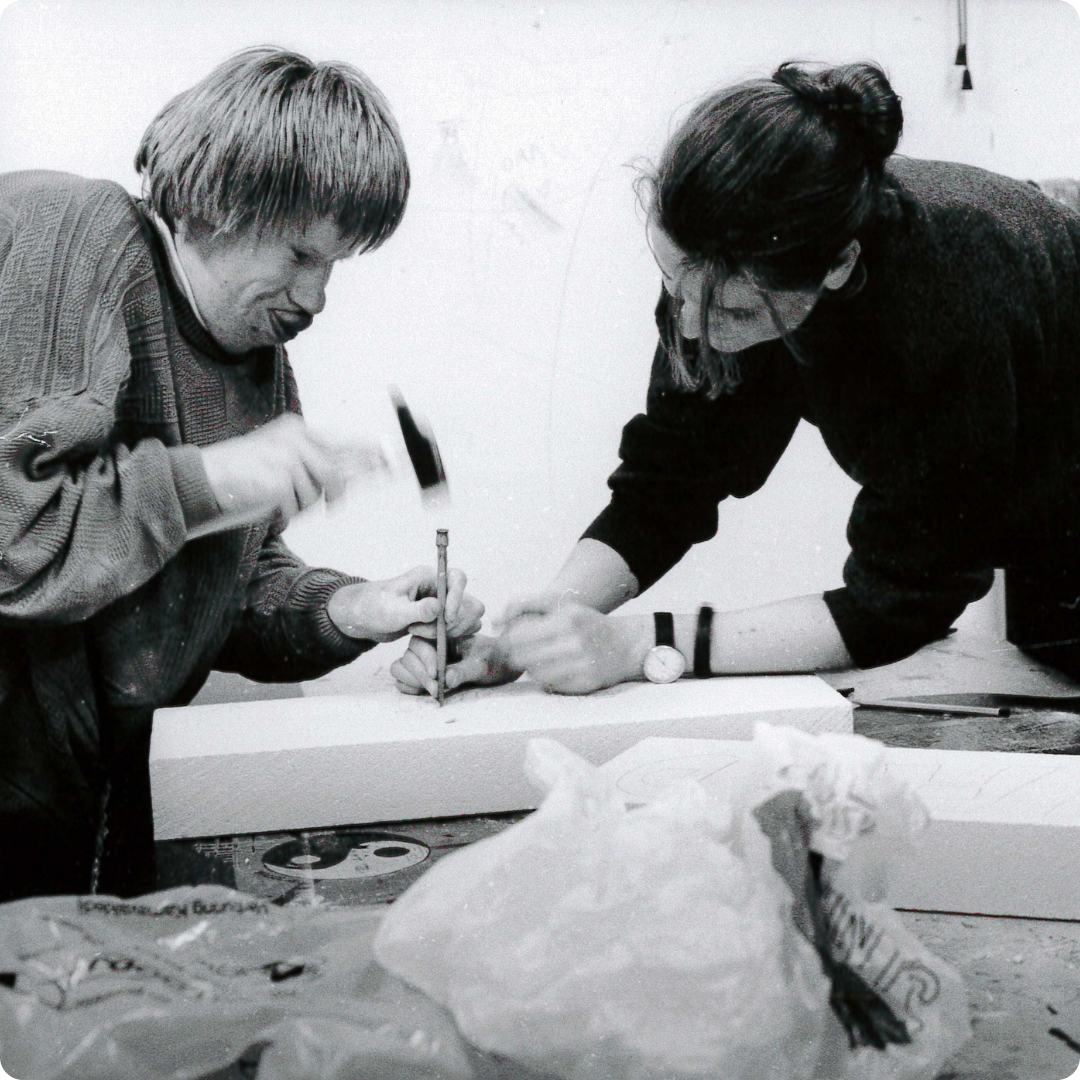 Man en vrouwelijke begeleidster kloppen met hamer een nagel in een plank. Foto in zwart wit.