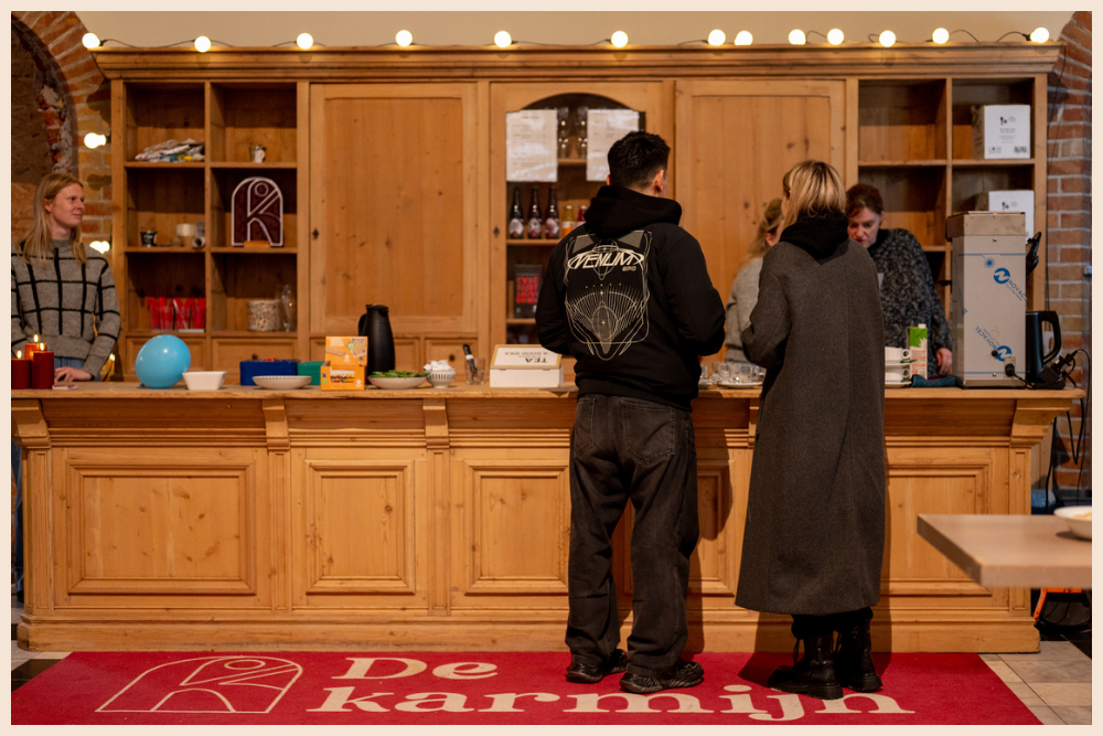 "Interieur van café 'De Karmijn' met een grote houten toog, personeel en klanten, en een rode vloermat met logo van De Karmijn.