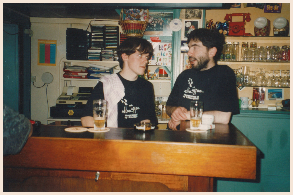 Twee mannen in zwarte T-shirts in gesprek achter de bar van café 'Alloo Alloo', met bierglazen op de toog