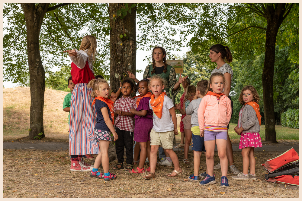 Een groep kinderen met oranje sjaaltjes luistert buiten in een bosrijke omgeving naar een begeleider in een traditioneel kostuum