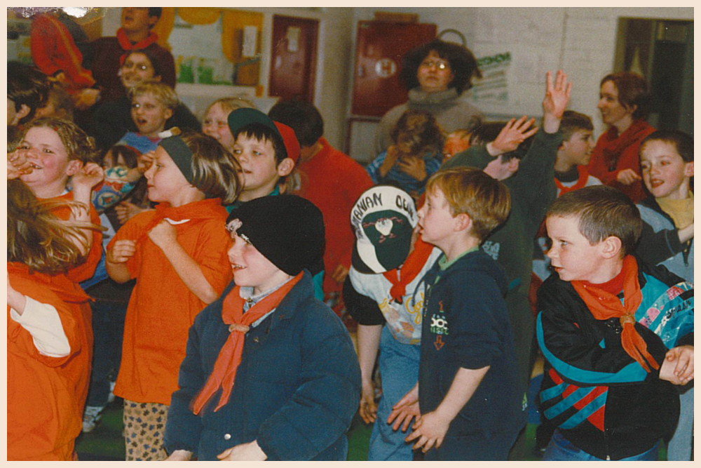 Een binnenopname van een groep kinderen, waarvan velen oranje sjaaltjes dragen, tijdens een vrolijke activiteit.