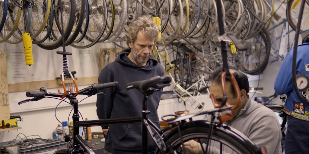 Man kijkt naar andere man die aan een fiets aan het sleutelen is, al lerend.